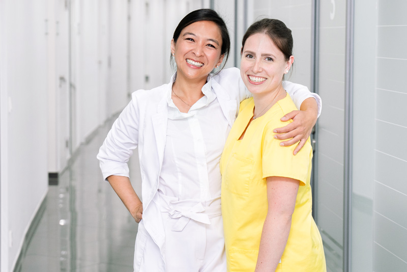 Zahnärztin steht mit ihrer Assistenz im Gang der Praxis, beide lächeln in die Kamera